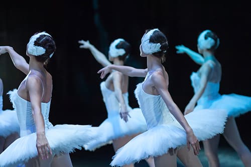 ballet performers stretch out their arms while on stage with a blue light shining on them